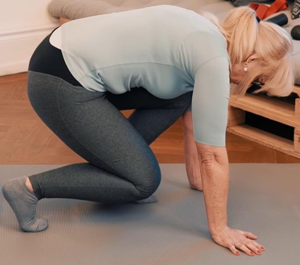 A woman does a stretching excercise, looks like she tries to stand up from her knees after sitting on the floor. She put her hands in front of her in order to lift herself up.