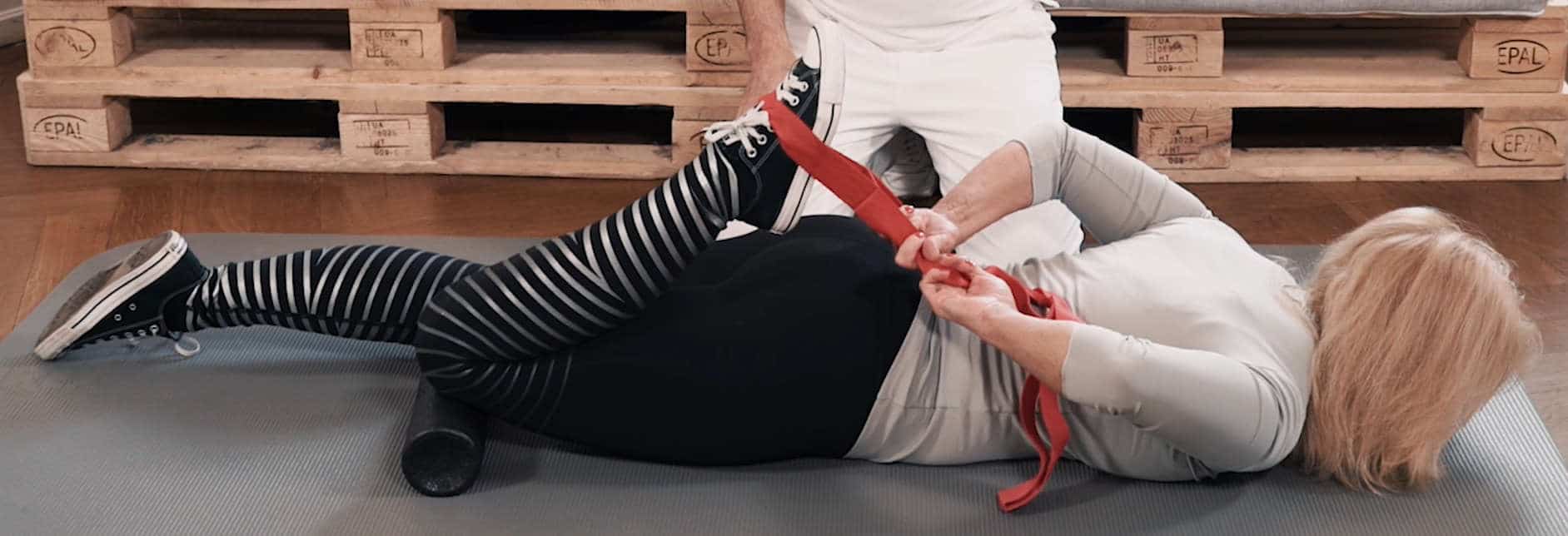 A woman lies on the floor with her face down. Using a stretching strap she pulls one leg bent at the knee towards her body to stretch the muscles. A man sits behind her.
