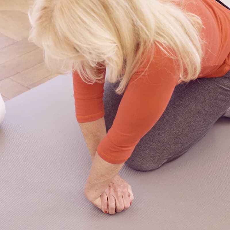 A woman sits on her knees and leans on her straight hands in front of her. Right hand lies over the left hand.