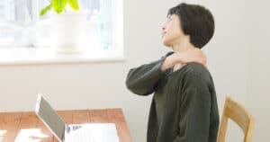 A woman is sitting at a desk in front of a laptop. She is rubbing her aching frozen shoulder with her eyes closed.