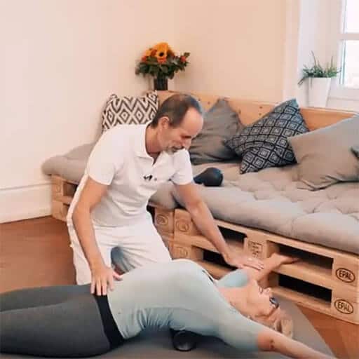 A woman lies on her back bending over a midi foam roller, which is placed under her shoulder blades. She spreads her straight arms above her head. A man, Roland Liebscher-Bracht sits next to hear holding his arms on her belly and right arm.