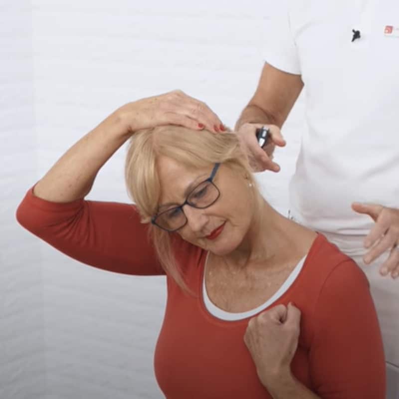 A woman tilts her head to the right towards the shoulder. She pulls her head further to the right with the help of her right hand. She bends her left arm in an elbow and holds her fist next to her chest.
