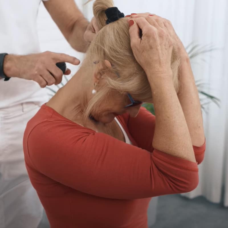 A woman holds her hands on her head and pulls it forward to stretch the neck muscles. A man stands behind her and points with one finger at one of her neck vertebrae.