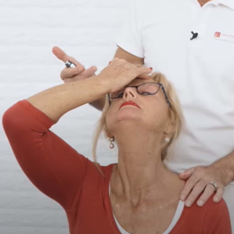 A woman throws her head backward with closed eyes. She holds her right hand on her forehead. A man stands behind her placing his arms on woman's shoulders.