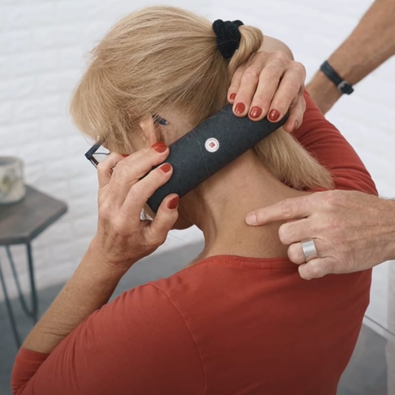 A woman holds a mini foam roller in both hands and rolls it along the back line of her neck. A man points with his finger at one particular spot on her shoulder.