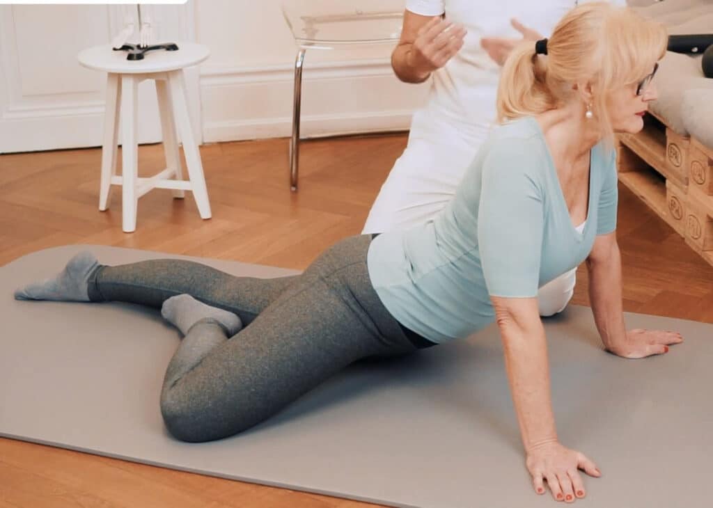 A woman lies face down on the floor leaning on her straightened hands. Right leg is bent at the knee and placed on the right from the body . A man sits behind her on his knees and explains the exercise by articulating with his hands.