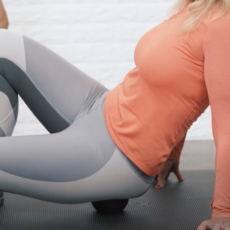 A woman in sportswear places her buttock on a midi massage ball. She does the exercise on a grey mat on the ground.
