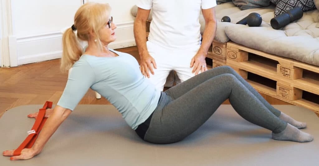 Ina sits on a mat. Her hands hold a stretching strap with her arms straightened and wide apart. Her legs are slightly bent; her feet touch the ground.
