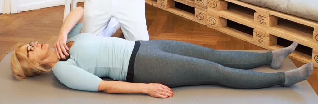 A woman is lying on a mat, massaging her shoulder with a massage ball.