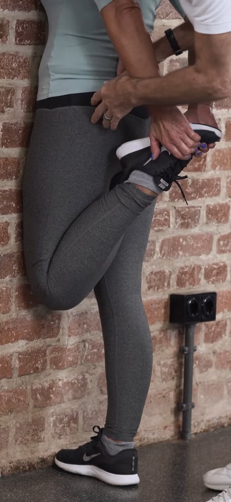 A woman is standing at a wall, one leg pulled up to stretch.