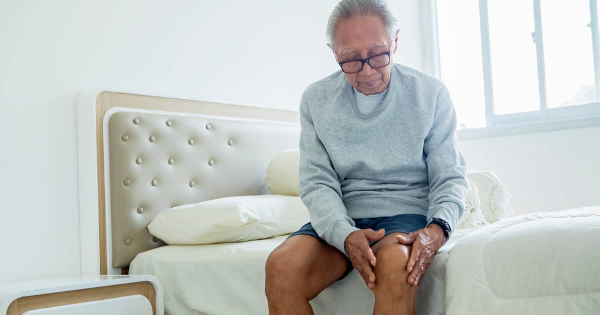 An old man is sitting on the edge of a bed, holding his hurting knee.