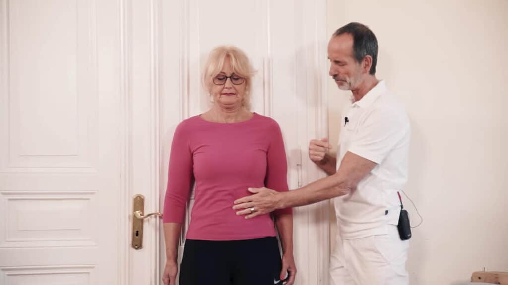 A woman stands straight up with closed eyes and deeply inhales. She leans on the white door with the head, shoulders, back and hips flat against the wall. A man, Roland Liebscher-Bracht stands next to her and holds his left hand on her stomach.