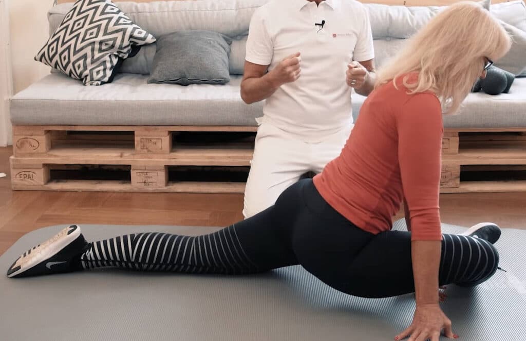 A woman lies on the floor lifting her upper body with straightened arms. She bends her right leg in front of her at a ninety degree angle. A man, Roland Liebscher-Bracht sits behind her on his knees and explains the exercises with his hands.