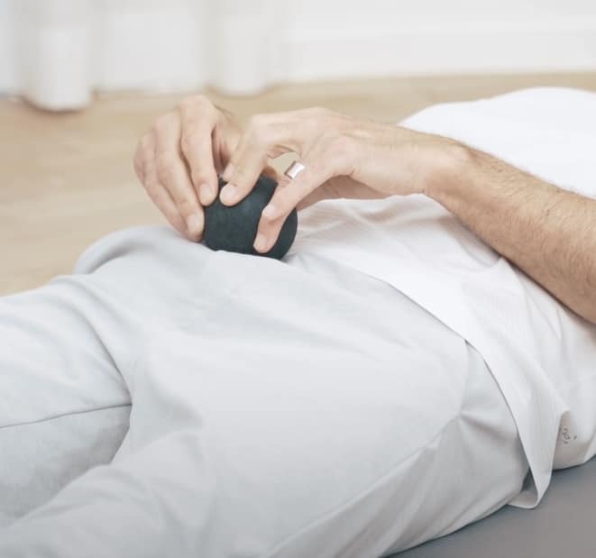 Two hands hold a massage ball that is pressed into the right side of the hip. Thighs, trunk, hands and arms are visible. The person lies on the ground.