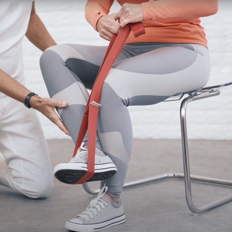 Ina sits on a stool. Her left foot touches the ground. Her right foot is attached to a stretching strap, her hands pull the foot towards her upper body.