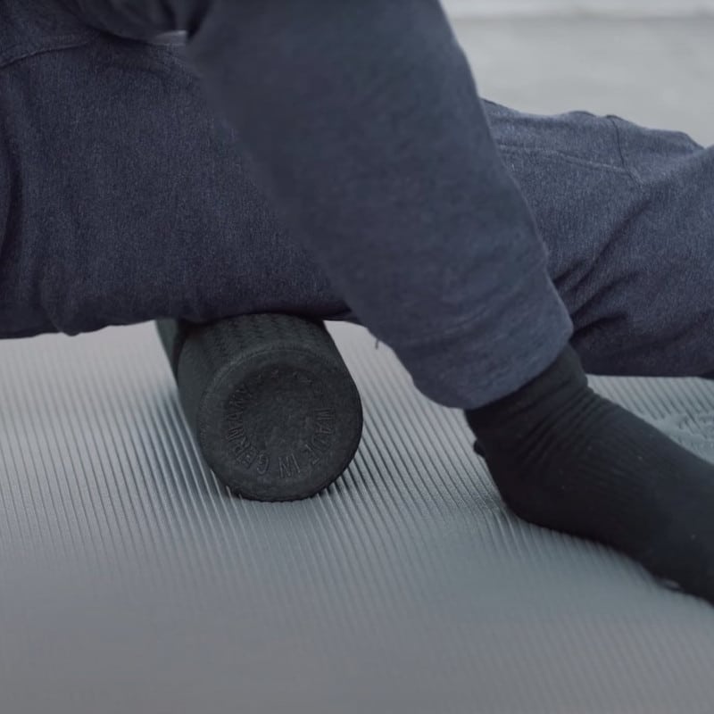 Roland Liebscher-Bracht sits on the ground and rolls over a midi foam roller that is placed under his right thigh. The left leg is bent, and both arms are straightened.