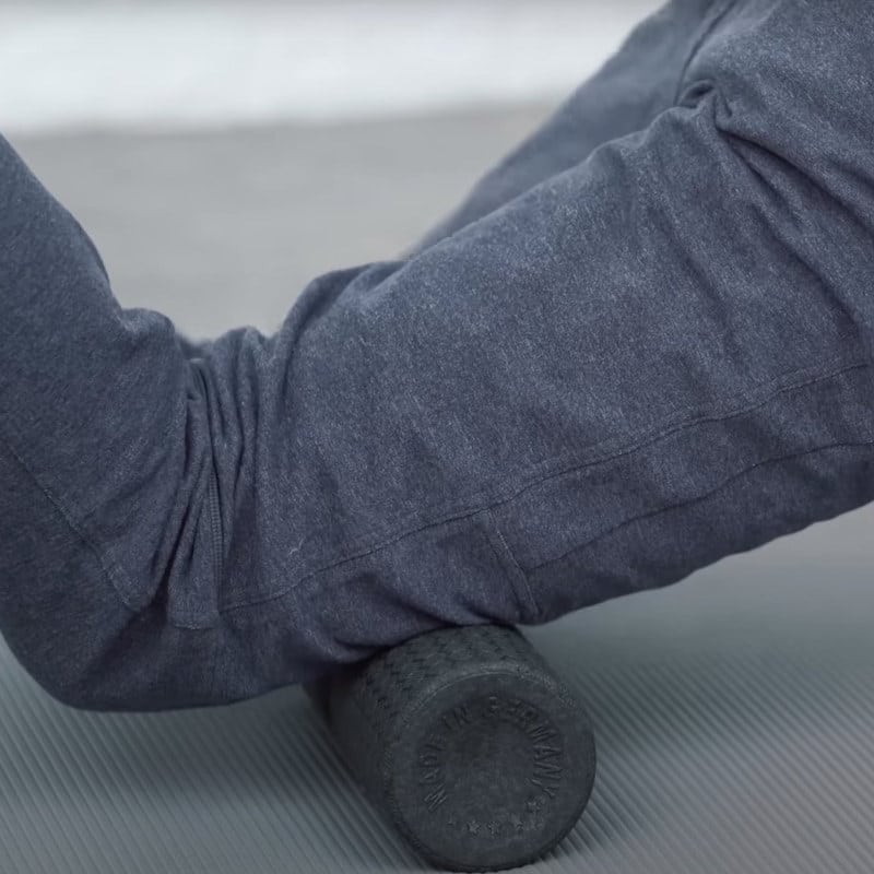 Two thighs balance on a midi foam roller that is placed on a mat. The person wears blue-ish sportswear.