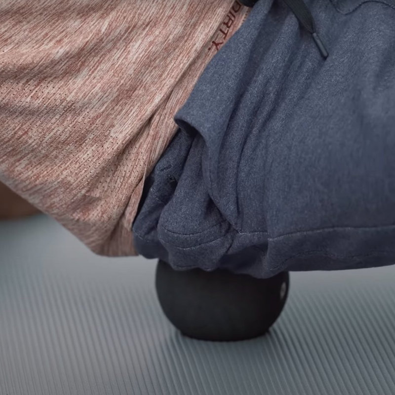 A person balances their buttocks on a black massage ball that is placed on a mat. Only the ball, hip and a part of thighs and stomach are visible.