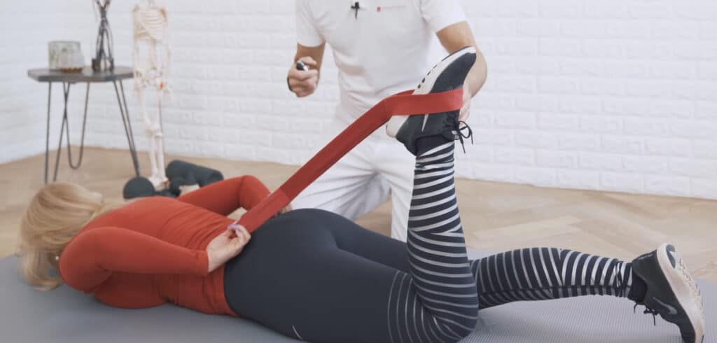 A woman is lying on the ground, face-down. She is tugging her leg towards her buttocks using a stretching strap as help.