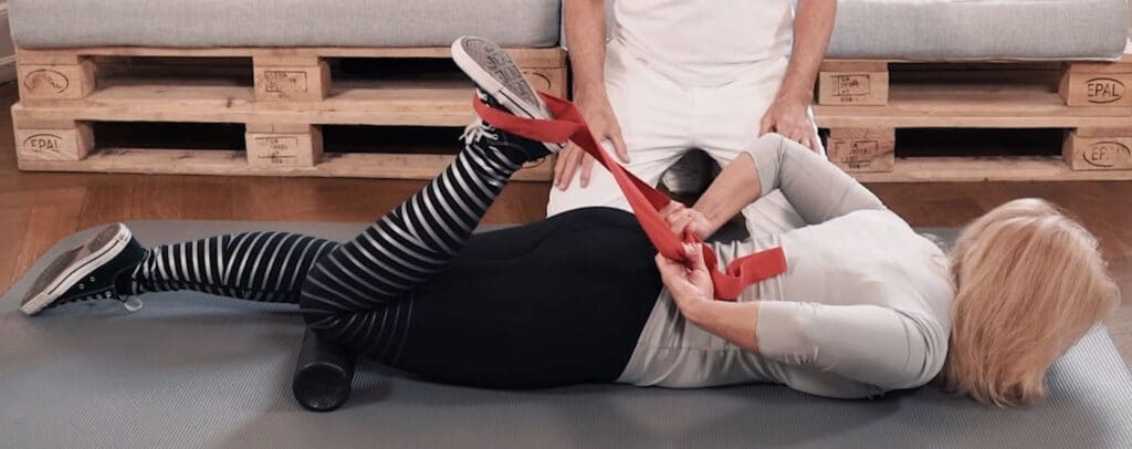 A woman lies face down on the floor and bends her left leg at the knee. She pulls her heel towards her buttlocks using red stretching strap. A mini foam roller is placed under her knee to intensify the stretching. A man sits on his knees behind her holding his palms on his legs.