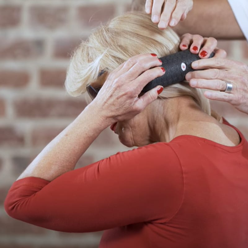 Ina holds a mini foam roller with both hands. The foam roller touches the left side of her neck. Two other hands are visible.
