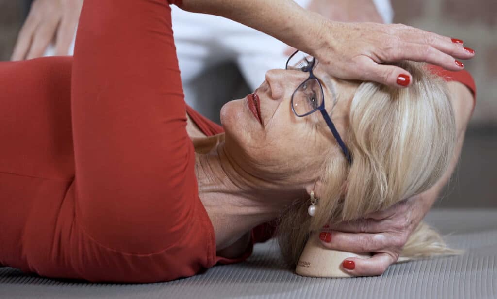 A woman is lying on the ground, pressing a point behind her ear with an osteopressure tool.