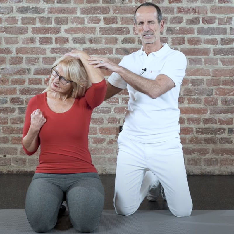 Ina kneels on the ground. She stretches her neck by pulling her head with the left hand towards the ground. The right hand forms a fist, the right shoulder pushes downward. Roland Liebscher-Bracht kneels next to her.