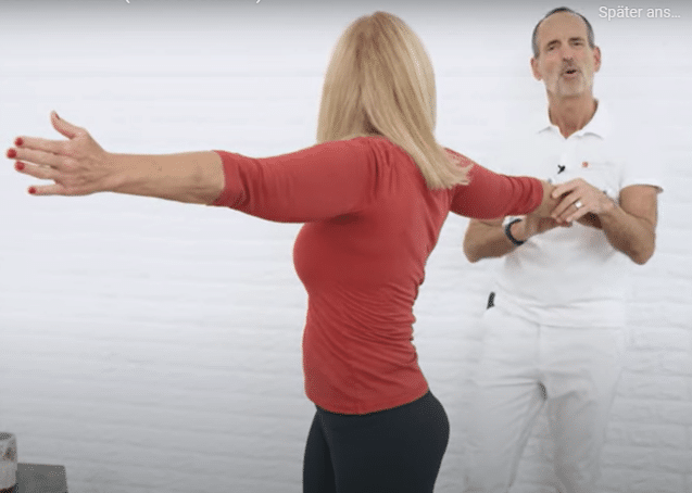 A woman is stretching her arms straight out, she's twisting her upper body to the right while Roland Liebscher-Bracht looks on.