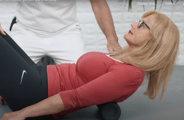 A woman is laying on the floor massaging her back with the Midi Foam Roller while Roland Liebscher-Bracht looks on.