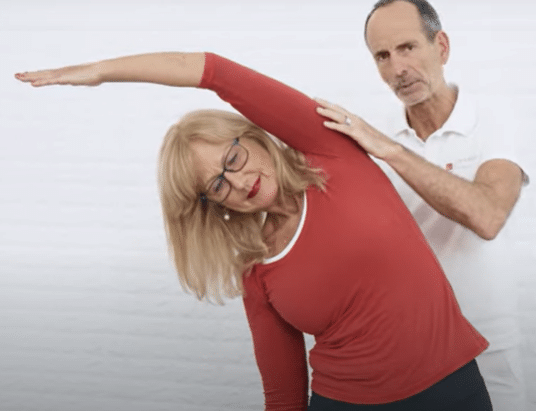 A woman is reaching to the left to stretch her ribcage while Roland Liebscher-Bracht looks on.