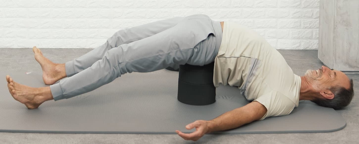Roland lies on the ground. A black stack is placed unter his sacrum and lift up the buttock. Shoulders, head, arms and feet touch the floor.