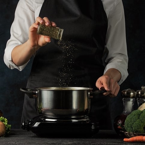 a chef in a black apron adding spices to a saucepan with soup