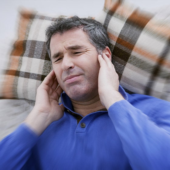 A man with tinnitus is holding his ears and creases his face in pain. Two chequered pillows are arranged behind him.