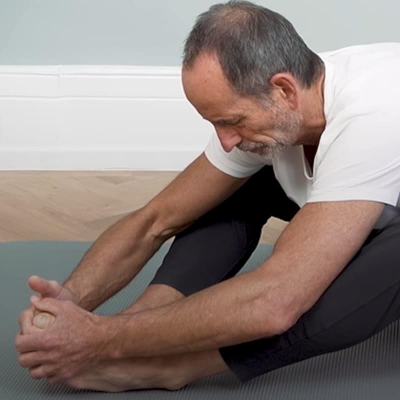 Roland Liebscher-Bracht is sitting on the floor, his legs are slightly bent, the soles of his feet touching his other. His hands are touching his feet and he is pulling forward to stretch his body.