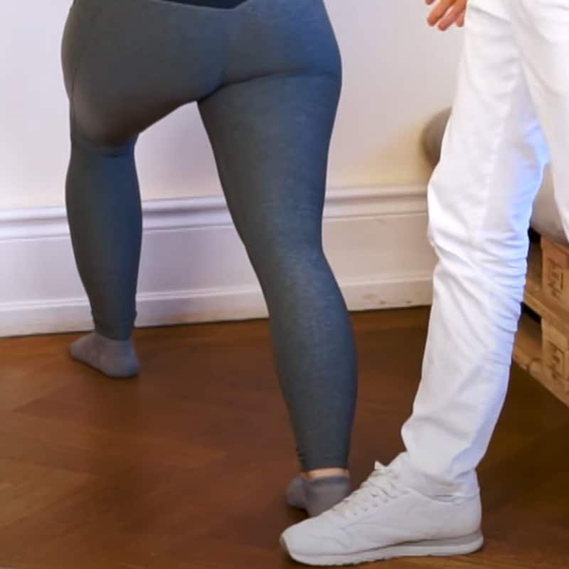 A woman is standing with the toes of her right foot against a wall, her left foot is stretched back.