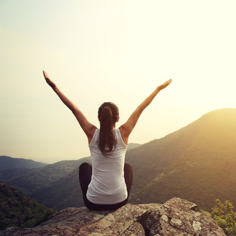 A girl is sitting on a cliff, stretching out her arms as part of a chest and back workout.