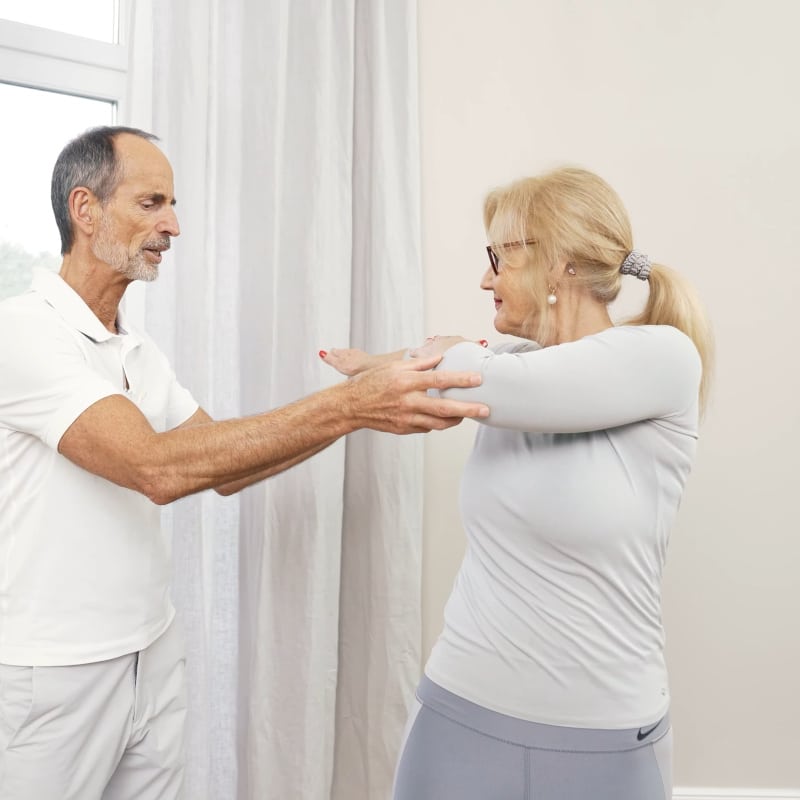 Ina's arms are raised horizontal to her shoulders and Roland Liebscher-Bracht is helping her rotate her upper body to the right.