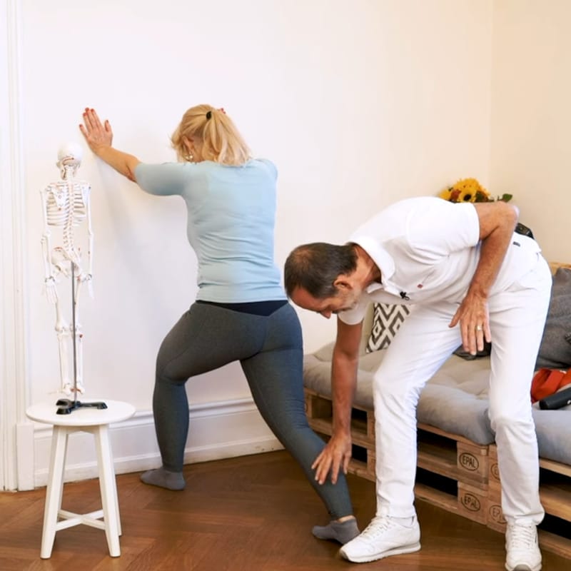 Roland Liebscher-Bracht is touching a woman's calf as she demonstrates a wall stretch for heel pain.