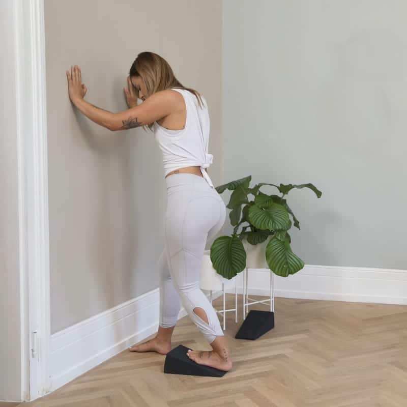 A woman is stretching her left calf. She's standing in front of a wall, the toes of her right foot are against the wall and her left foot is on the Knee Hero.