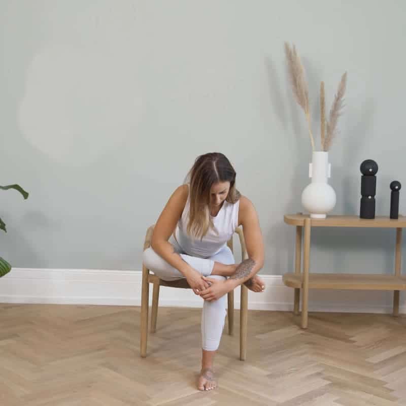 A woman is sitting in a chair. Her left ankle is resting on her right knee, she's leaning forward stretching her buttocks.