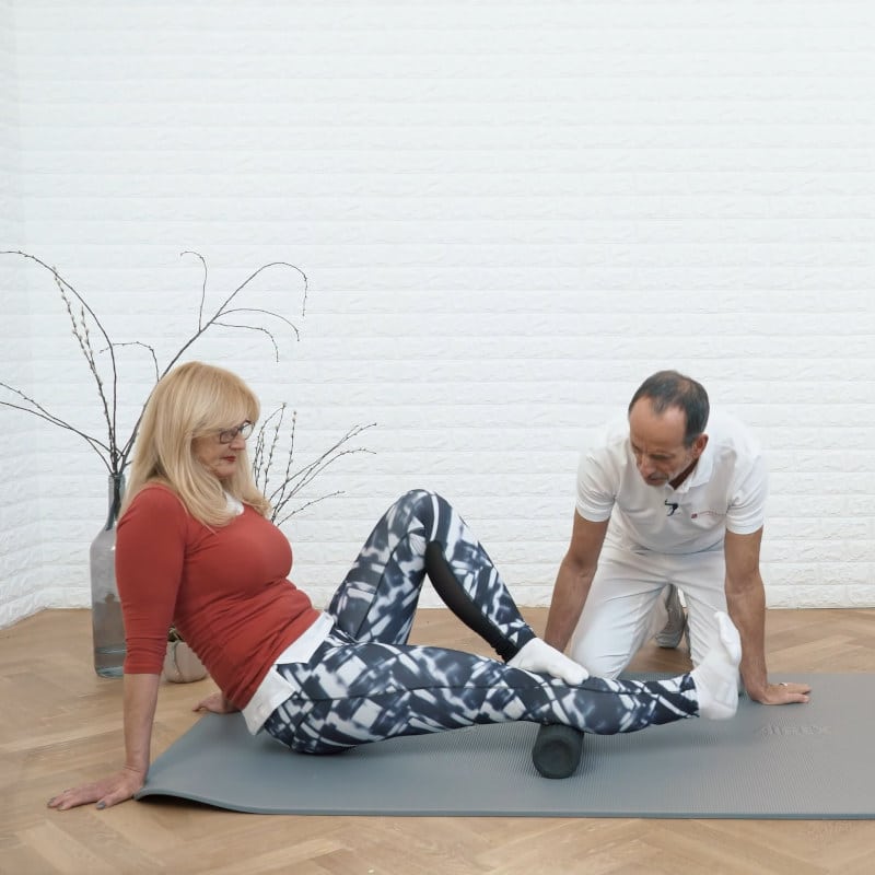 A woman is sitting on the floor foam rolling the back of her leg while Roland Liebscher-Bracht looks on.