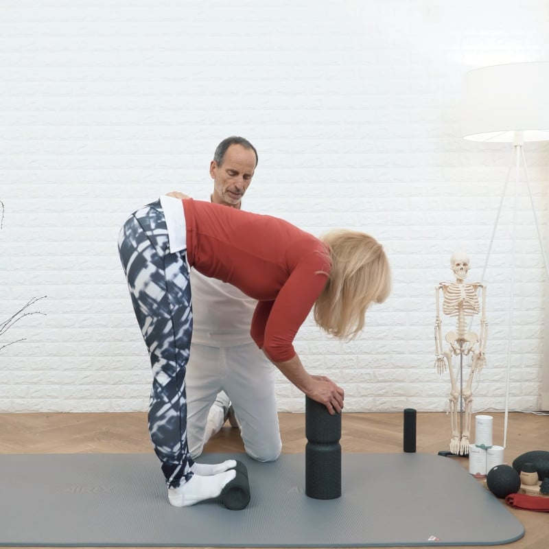 A woman is standing with the midi foam roller under her toes and she's leaning forward with her hands on the maxi foam roller as Roland Liebscher-Bracht looks on.