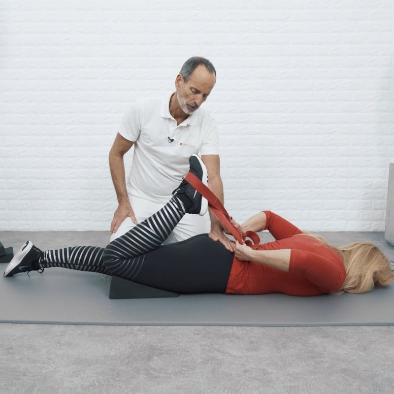A woman is demonstrating a knee & thigh stretch for Osgood Schlatter. She is laying on her stomach with the Knee Hero wedged under her thigh, the Stretching Strap is around her foot. Roland Liebscher-Bracht is checking her form.