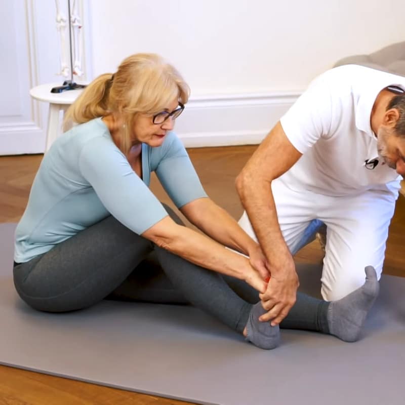 A woman is sitting on the floor with her right leg bent slightly. Roland Liebscher-Bracht is touching her foot..
