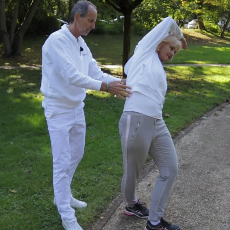 A woman is bending her upper body to the right, stretching her hip while Roland Liebscher-Bracht looks on.