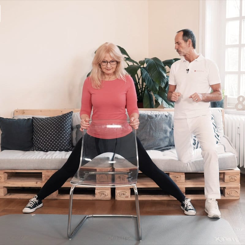 A woman is standing behind a chair assuming a wide stance as Roland Liebscher-Bracht looks on.