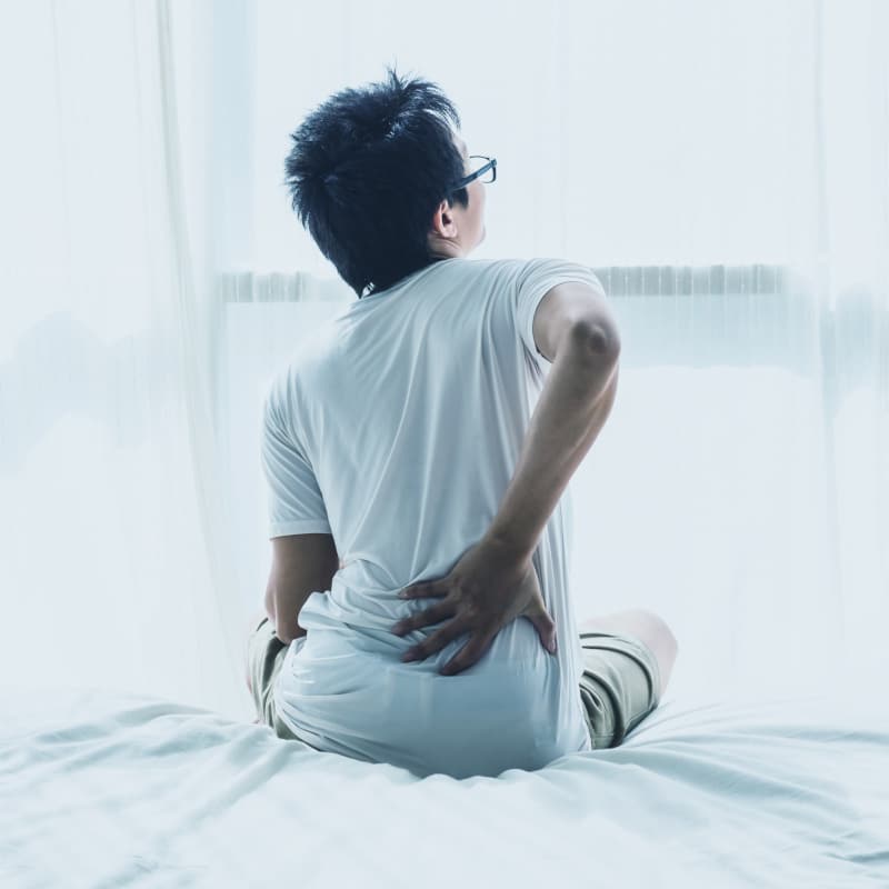 Closeup of a woman sitting on a bed, she's touching her lower back.