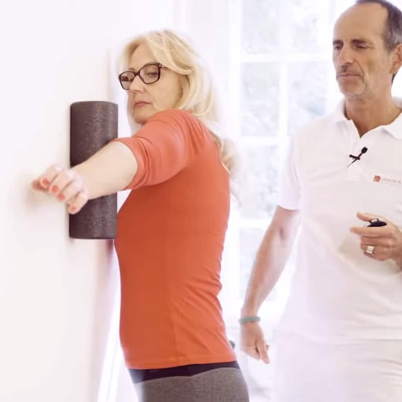 A woman is foam rolling the inside of her elbow against a wall.