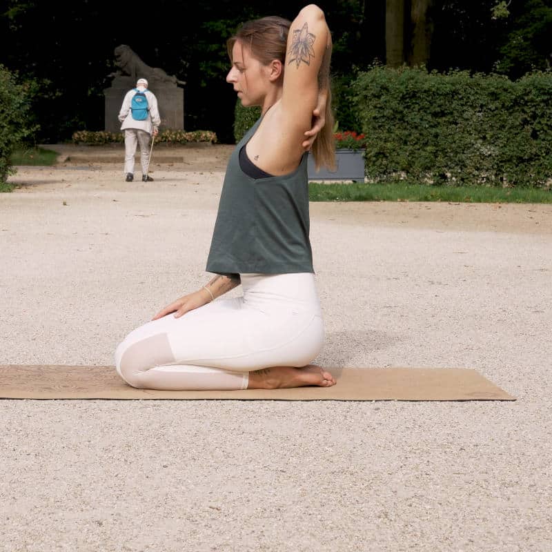 A woman is sitting back on her heels reaching back with her right hand touching her shoulder.