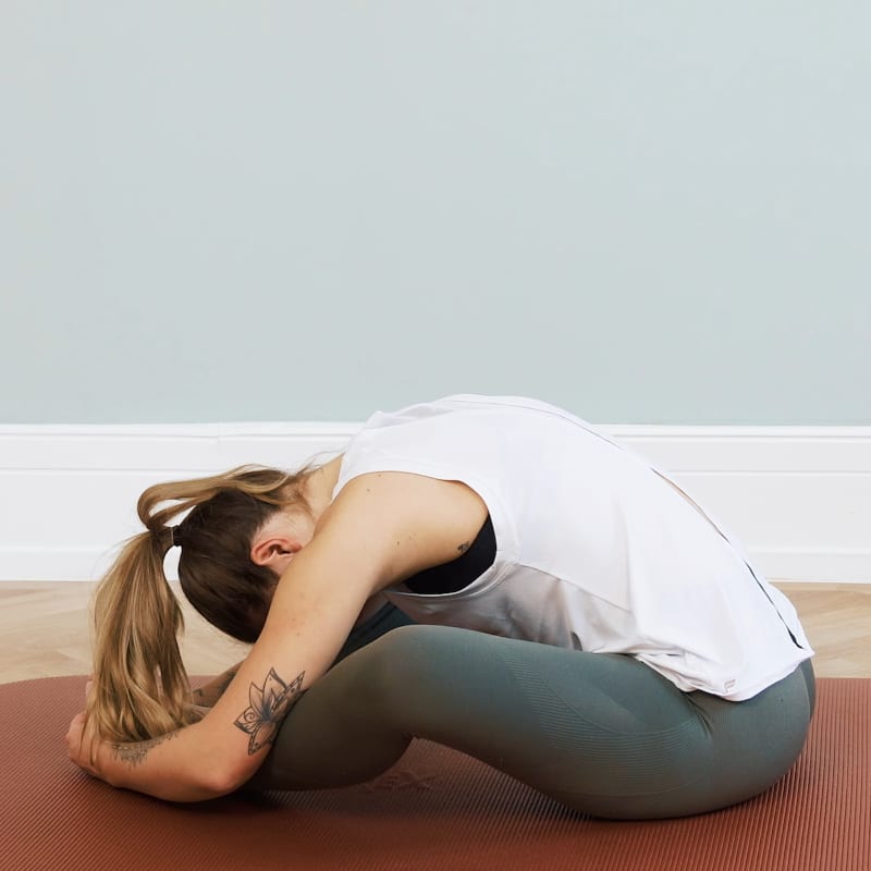 A young woman is sitting on the floor, she's grabbing her feet and pulling her upper body forward.